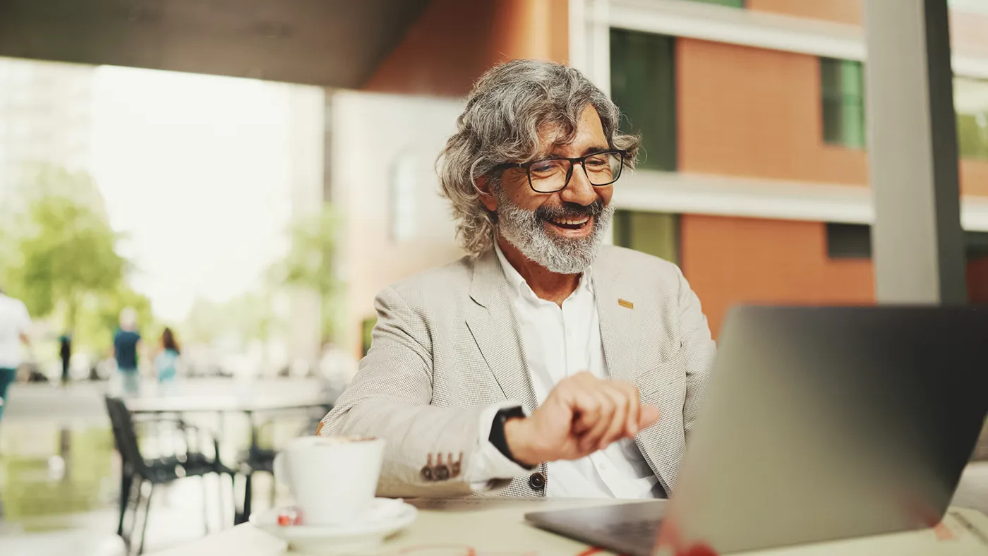 An older man with a gray beard and glasses smiles as he works on a laptop in an outdoor cafe setting. He is dressed in a light-colored blazer, and a cup of coffee sits on the table beside him, adding to the relaxed and productive atmosphere. The blurred background of trees and modern architecture suggests a pleasant urban environment.