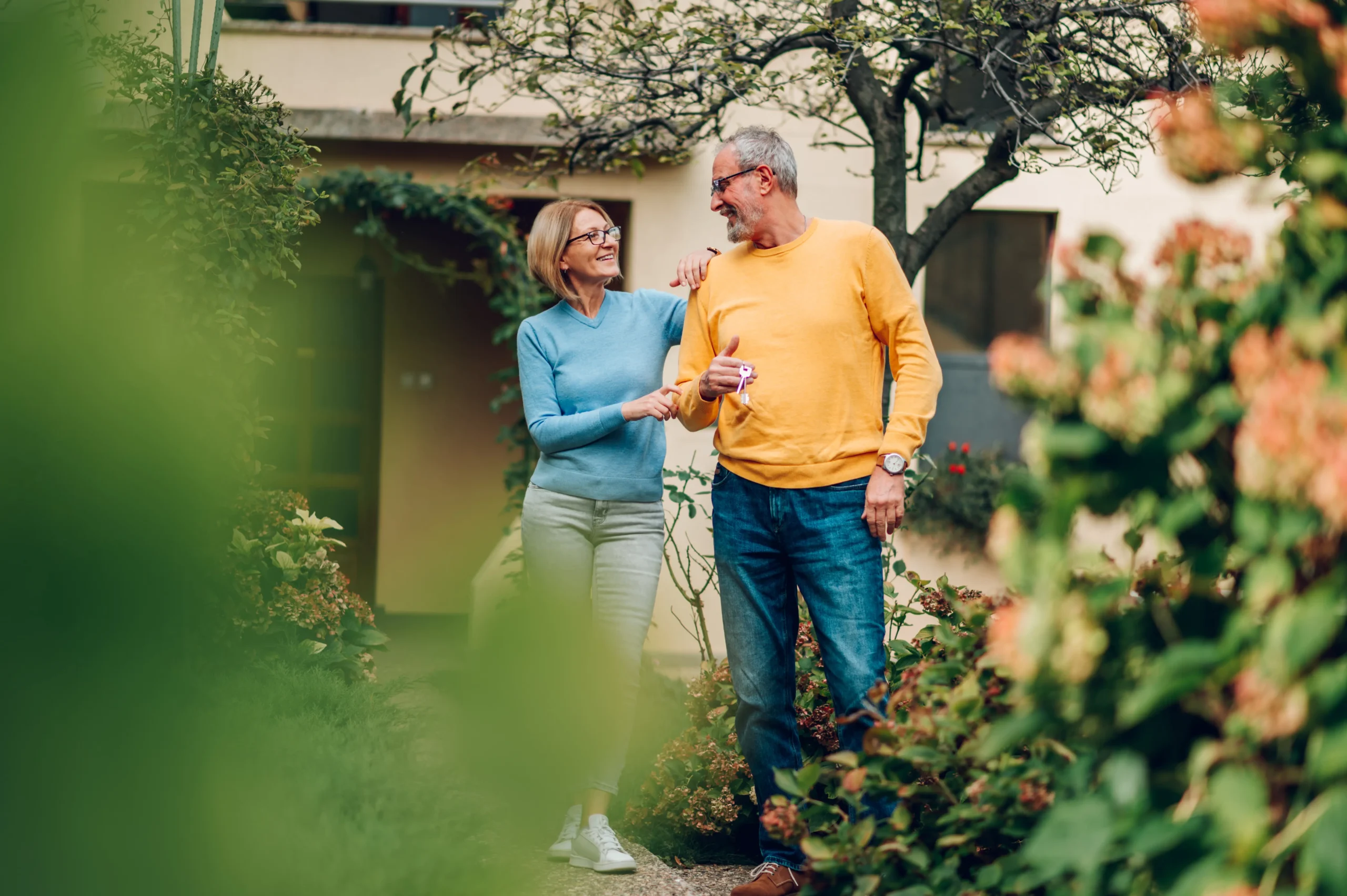 A happy senior couple walks together in a lush garden, with the man holding a set of keys and smiling at his partner. The woman affectionately holds his arm, both dressed casually in bright, contrasting sweaters. The natural surroundings and their joyful expressions create a warm, peaceful atmosphere.